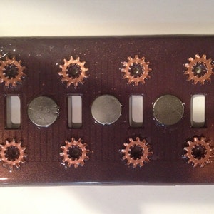 May include: A dark brown light switch cover with a steampunk design. The cover features eight copper gears and three silver metal circles. The cover has a gradient from dark brown to light brown.