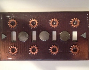 May include: A dark brown light switch cover with a steampunk design. The cover features eight copper gears and three silver metal circles. The cover has a gradient from dark brown to light brown.