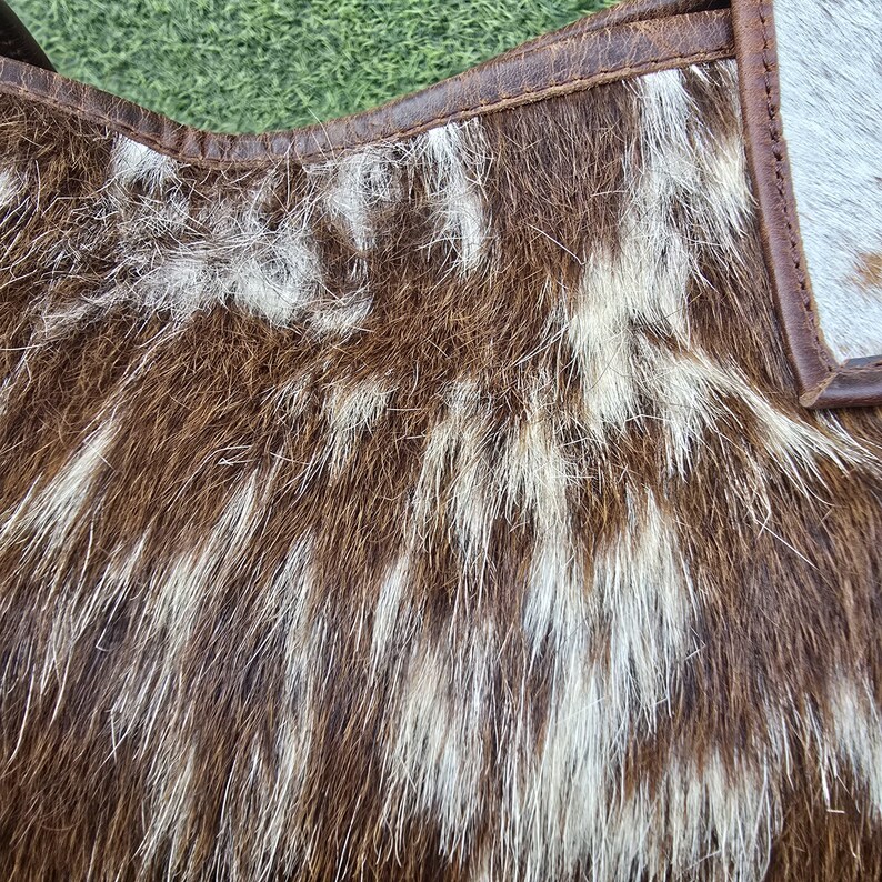 May include: Close-up of a handbag made from brown and white cowhide. The bag features a natural, textured pattern with a mix of dark brown and creamy white fur. The edges are trimmed with brown leather.