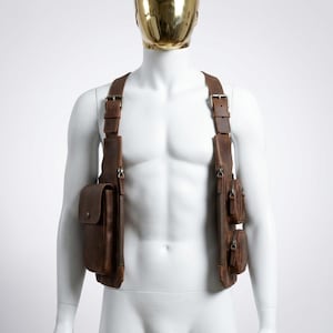 May include: A brown leather chest harness featuring two vertical panels connected by adjustable shoulder straps with metal buckles. The right side includes a large rectangular pocket with a snap closure, while the left side features two smaller circular zippered pouches.