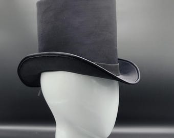 Puede incluir: Un sombrero de copa negro con una corona cilíndrica y un ala ancha y curvada. Una banda negra rodea la base de la corona. El sombrero se exhibe sobre una cabeza de maniquí blanca.