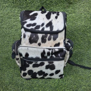 Puede incluir: Mochila con estampado de piel de vaca en blanco y negro. La bolsa tiene un cierre de solapa, un bolsillo frontal con cremallera y dos bolsillos laterales. La mochila tiene cremalleras y correas negras.