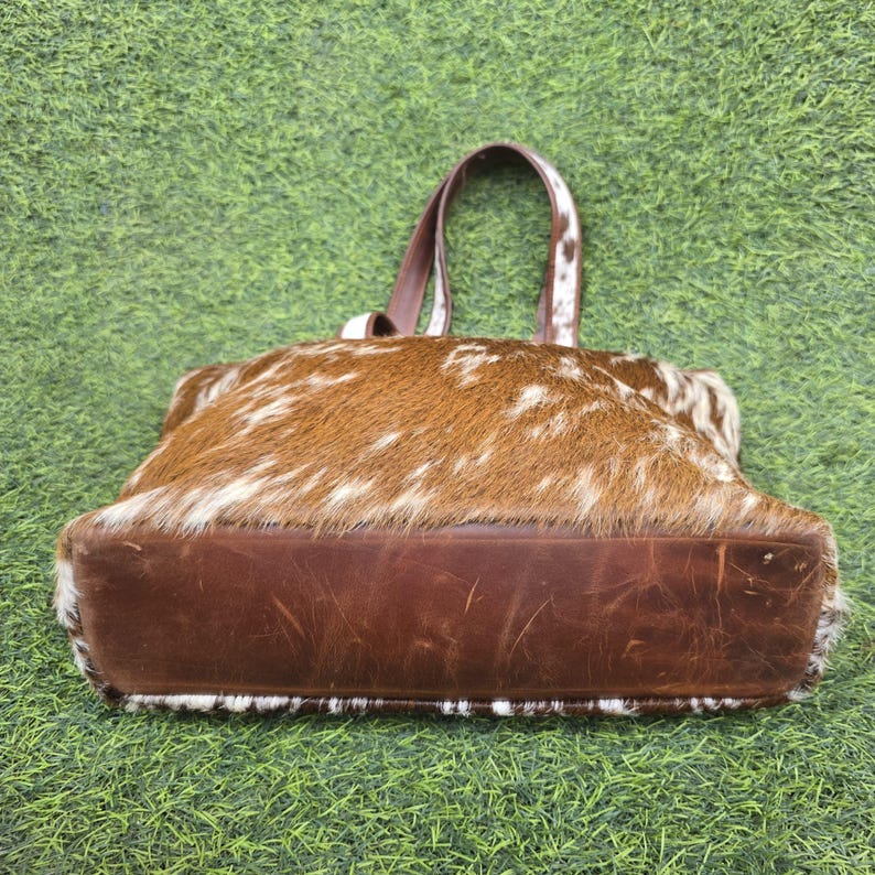 May include: A brown and white cowhide tote bag with a dark brown leather base and handles. The bag is rectangular with a rounded top and a natural, textured appearance. The handles are attached to the top of the bag.