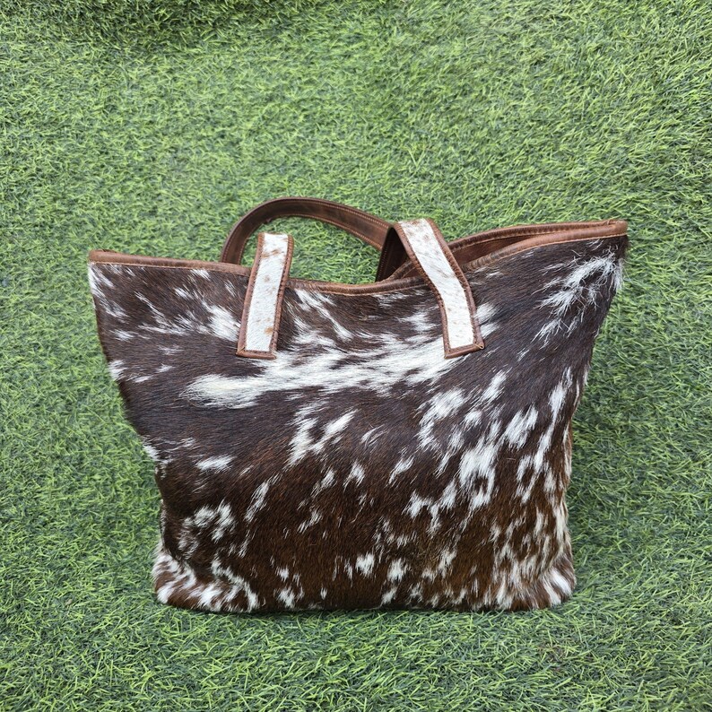 May include: A cowhide tote bag with a brown and white spotted pattern. The bag has brown leather handles and trim, and white accents on the handles. The bag is sitting on a green surface.