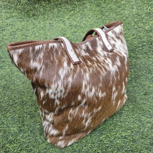 May include: A large tote bag made of brown and white cowhide. The bag has a rectangular shape with a wide opening and two sturdy handles. The exterior features a natural, spotted pattern, and the interior is lined with a brown material.