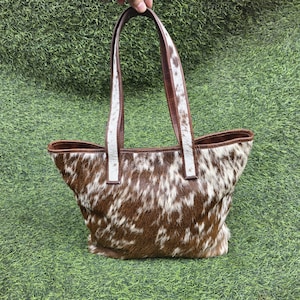 May include: A brown and white cowhide tote bag with brown leather handles and trim. The bag has a spacious interior and a natural, textured appearance. The handles are held by a hand against a green background.