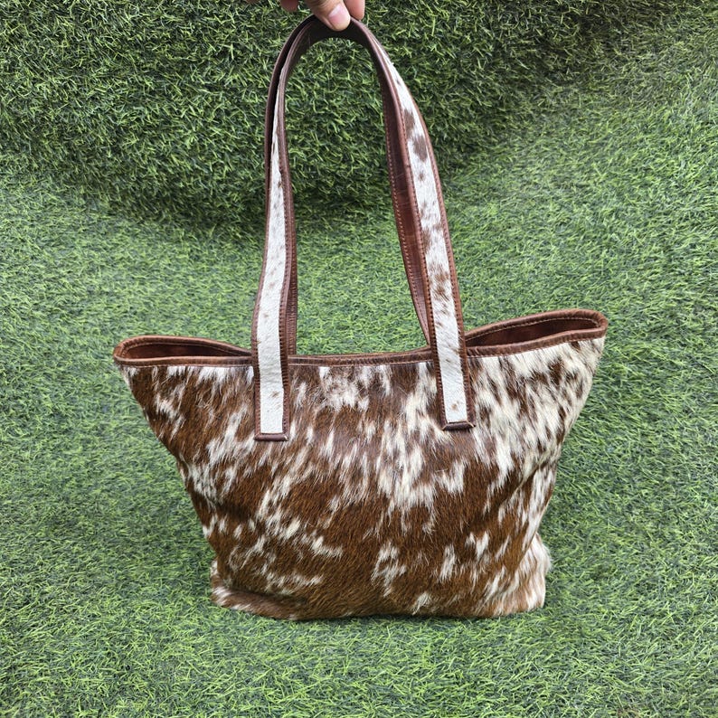 May include: A brown and white cowhide tote bag with brown leather handles and trim. The bag has a spacious interior and a natural, textured appearance. The handles are held by a hand against a green background.