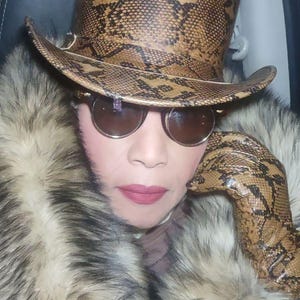 May include: A brown snakeskin-effect top hat and matching gloves are visible. The person is wearing round sunglasses and a fur coat. The hat has a wide brim and a tall crown. The gloves are long and reach up to the wrist.