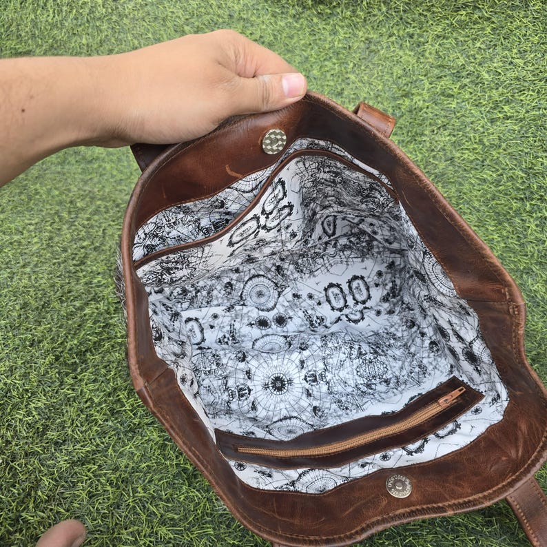 May include: A brown leather tote bag with a patterned interior. The bag has a zipper pocket and a button closure. The interior lining features a black and white design. The bag is held by a hand.