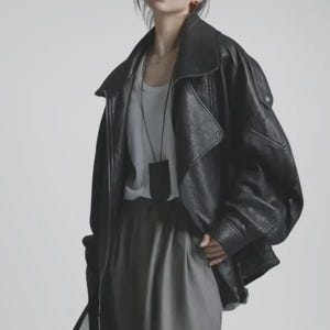 Puede incluir: Chaqueta de cuero negra con cuello ancho y corte holgado. La chaqueta se combina con una camiseta blanca y pantalones grises. Una pequeña bolsa negra cuelga de un collar.
