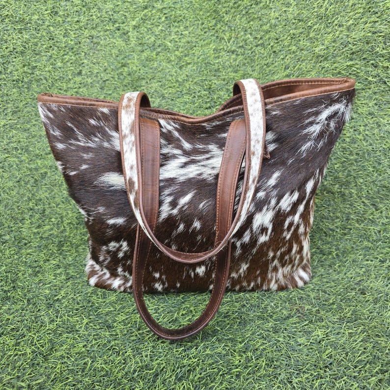 May include: A cowhide tote bag with a brown and white spotted pattern. The bag has brown leather handles and trim. The bag is sitting on a green surface.