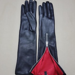 May include: A pair of black leather gloves with a long design. The gloves have a zipper detail on the side, revealing a red interior lining. The gloves are displayed on a neutral background.