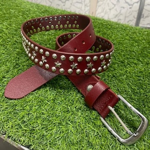Op de afbeelding: Een donkerrode riem voorzien van een reeks zilverkleurige metalen ster- en cirkelvormige studs over de lengte van de band. De riem is uitgerust met een zilverkleurige metalen gesp en een bijpassende rode riemlus, met een puntig uiteinde.