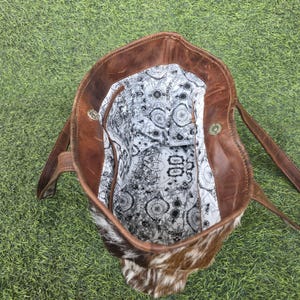 May include: A brown leather tote bag with a cowhide front. The bag has two handles and a patterned interior lining. The bag is open, revealing the interior lining with a map-like design. The bag is sitting on a green surface.