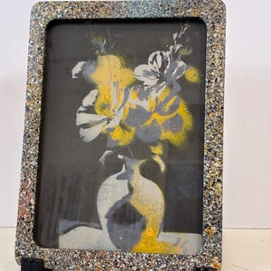 May include: A rectangular picture frame with a speckled, multi-colored granite-like texture. Inside, a print features a stylized vase containing yellow and grey flowers against a solid black background. The frame rests on two small black stands.