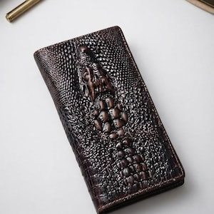 Puede incluir: Una cartera rectangular de color marrón oscuro con una textura en relieve que imita la piel de cocodrilo. La superficie muestra un patrón detallado de cabeza y lomo de cocodrilo en relieve con sombras profundas y acabado brillante.