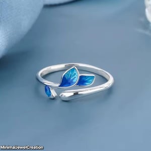 May include: A silver ring with a leaf design. The ring features three blue enamel leaves. The ring is open and adjustable. The text "MinimalJewelCreation" is at the bottom left.