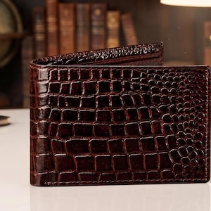 Può includere: Un portafoglio bifold marrone scuro caratterizzato da una finitura lucida e testurizzata che imita l'aspetto della pelle di coccodrillo. Il portafoglio ha una forma rettangolare con cuciture visibili lungo i bordi e un motivo a scaglie.