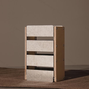 以下が含まれることがあります： 5つの水平な灰色の棚が付いた長方形の木製収納ボックス。ボックスは明るい茶色のフレームで、小さな金属ネジで固定されています。ボックスは茶色の表面に置かれています。