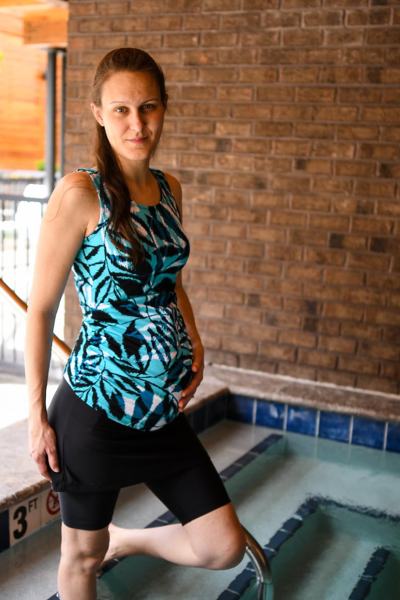 Puede incluir: Una persona embarazada lleva un traje de ba&ntilde;o estampado en azul y negro con una falda negra sobre la parte inferior. La persona est&aacute; de pie junto a una piscina con la mano en el est&oacute;mago.