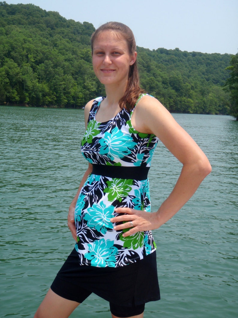 Puede incluir: Una mujer lleva un traje de ba&ntilde;o de estampado floral negro y azul con una falda negra. El traje de ba&ntilde;o tiene un cintur&oacute;n negro y un estampado floral verde y azul. La mujer est&aacute; de pie frente a una masa de agua.