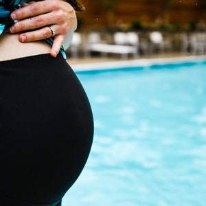 Puede incluir: Una mujer embarazada con un traje de ba&ntilde;o negro est&aacute; de pie junto a una piscina. Sus manos descansan sobre su vientre.