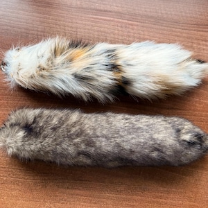 May include: Two faux fur tails, one in shades of grey and brown, the other in a mix of white, brown, and black. The tails are fluffy and soft, with a small metal loop at the base. They are laid out on a wooden surface.