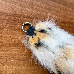 May include: A keychain featuring a fluffy, spotted fur tail with a gold-colored metal ring and a black leather loop. The fur is primarily white with patches of black and tan.
