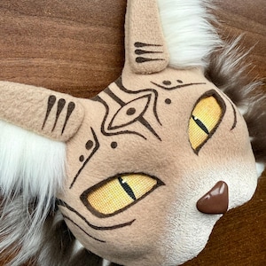 Può includere: Una maschera da gatto di pelliccia bianca e marrone con segni marroni e occhi gialli. La maschera ha un design tribale sul viso.