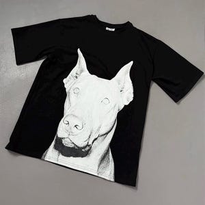 Può includere: Una t-shirt nera a maniche corte con una grande stampa grafica bianca ad alto contrasto raffigurante la testa di un cane Dobermann sul davanti. Il design mostra il muso del cane con orecchie a punta e tratti dettagliati sul petto.