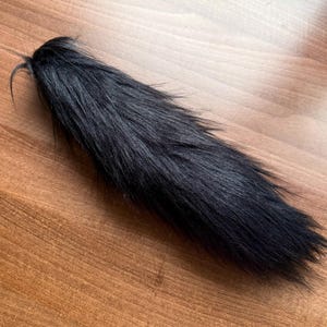 以下が含まれることがあります： 木製の表面に置かれた黒いフェイクファーの尻尾。 尻尾は長く先細りで、密度が高くふわふわした質感です。 尻尾の根元は集められ、固定されています。 コスプレやコスチュームに適しています。