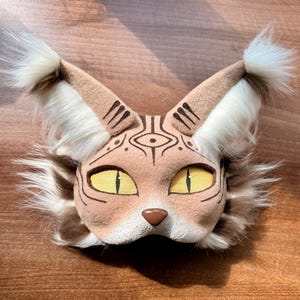 May include: A tan cat mask with large, pointed ears and yellow eyes. The mask features intricate brown markings around the eyes and on the ears. The ears and cheeks have fluffy white and brown fur accents. The nose is brown.