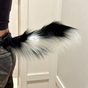 May include: A faux fur tail accessory featuring a long, bushy design with a colour pattern of black and white sections. The tail is attached to a black belt-like harness component that secures around the waist, displaying a soft, layered texture.