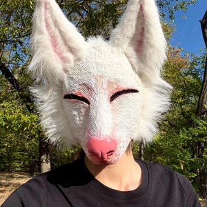 Op de afbeelding: Een wit vossenmasker met roze accenten op de neus, binnenoren en rond de ogen. Het masker heeft een textuur en pluizig haar. De persoon draagt een zwart t-shirt.