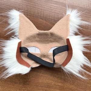 May include: A tan and white faux fur mask with pointed ears and eye cutouts. The mask has a brown trim and a black elastic band for securing it. The mask is designed to resemble a lynx or similar wild cat.