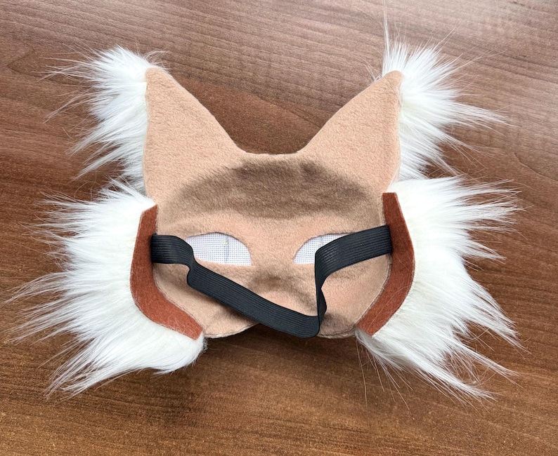 May include: A tan and white faux fur mask with pointed ears and eye cutouts. The mask has a brown trim and a black elastic band for securing it. The mask is designed to resemble a lynx or similar wild cat.