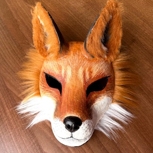 Op de afbeelding: Een handgemaakt vossenmasker met oranje, witte en zwarte details. Het masker heeft een realistisch ontwerp met een getextureerde, vachtachtige uitstraling rond de oren en het gezicht. De ogen zijn zwart en de neus is zwart.