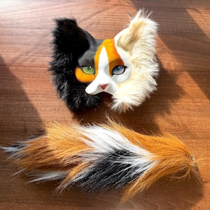 Può includere: Una maschera da gatto con pelliccia sintetica nera e crema, un volto dipinto con un occhio verde e uno blu, e una lunga coda in pelliccia sintetica abbinata con un motivo tricolore arancione, nero e bianco. La maschera ha un naso rosa.