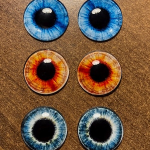Könnte beinhalten: Sechs Glas-Augen-Cabochons in verschiedenen Farben. Zwei sind blau, zwei sind orange und rot, und zwei sind blau und weiß. Jedes hat eine schwarze Pupille und einen weißen Glanz, ideal für Bastelarbeiten oder die Herstellung von Puppen.