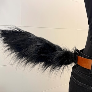 May include: A black faux fur tail attached to a belt. The tail is long and fluffy, with a slight curve. The belt is brown leather, and the jeans are black.