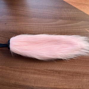 May include: A long, soft, light pink faux fur tail accessory. The tail features a tapered, pointed end and is attached to a short, black fabric loop at the base, which is secured with a small metal rivet for attachment or hanging purposes.