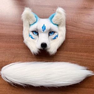 May include: A white and blue fox mask with matching fluffy tail. The mask features blue accents around the eyes and ears, with a flame design on the forehead. The tail is long and white, complementing the mask's design.