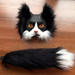 May include: A black and white cat mask and tail set. The mask features orange eyes, a pink nose, and white fur detailing. The tail is black with a white tip and a metal clasp for attachment.