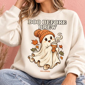 Op de afbeelding: Een crèmekleurig sweatshirt met ronde hals en een afbeelding van een cartoongeest met een oranje gebreide muts, die een dampende koffiebeker met een pompoenpictogram vasthoudt. De tekst luidt BOO BEFORE BREW en HALLOWEEN, omringd door herfstbladeren.