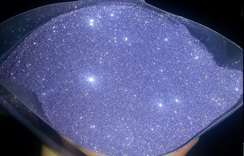 May include: A close-up view of a container filled with fine, iridescent purple glitter. The glitter is very sparkly and reflects light.