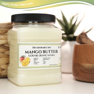 May include: A white plastic jar with a black lid labeled "Mango Butter 100% Pure Organic Natural" by Dr. Adorable Inc. The label also includes details about the botanical source, method, part, usage rate, and country of origin. The jar is sitting on a wooden surface.