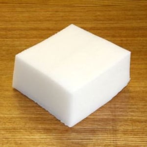 May include: A white rectangular block of solid soap on a wooden surface.