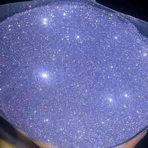 May include: A close-up view of a container filled with fine, iridescent purple glitter. The glitter is very sparkly and reflects light.