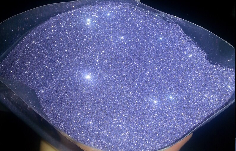 May include: A close-up view of a container filled with fine, iridescent purple glitter. The glitter is very sparkly and reflects light.