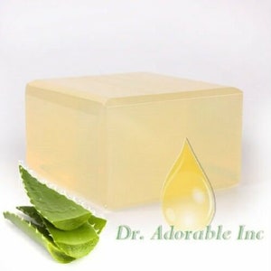May include: A clear, rectangular block of soap base with a yellow drop of oil on top. The soap base is sitting on a white surface with a green aloe vera plant in the foreground. The text "Dr. Adorable Inc." is visible in the lower right corner of the image.